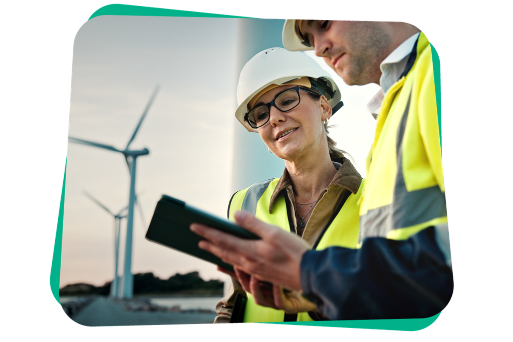 Two workers in safety gear discuss something on a tablet, with wind turbines in the background.