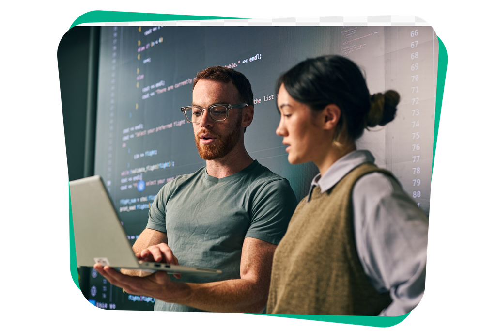 Two people discuss in front of a large screen displaying code, one holding a laptop.